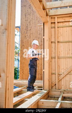 Carpenter constructing wooden framed house. Man worker cladding facade of house, using a screwdriver, wearing work overalls and helmet. Concept of modern eco-friendly construction. Stock Photo