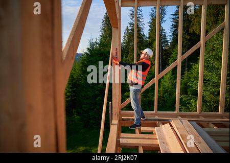 Carpenter constructing wooden frame house. Bearded man, wearing spectacles, inspects the walls for levelness using spirit level. Concept of environmentally-friendly contemporary construction. Stock Photo