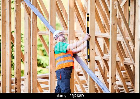 Carpenter constructing wooden frame house. Man inspects walls for levelness using spirit level, wearing protective overalls, helmet and vest. Concept of modern ecological construction. Stock Photo