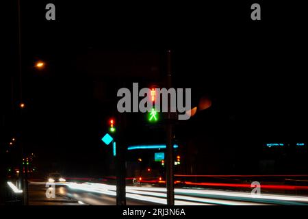 On a dark night, a row of cars drive past a traffic light with green and red colors, illuminated in the night sky Stock Photo