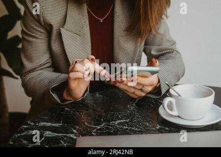 A young woman in her twenties is sitting at a table, looking down at her cellphone, seemingly engrossed in her phone activity Stock Photo