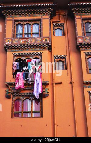 Colorful clothing hangs outside a bank of rabsel windows on an orange building with traditional painted decorations in Paro, Kingdom of Bhutan. Stock Photo
