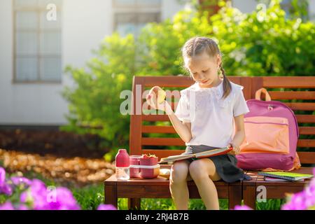 Pupil with lunch-box. Girl with backpacks is eating fruit near building outdoors. Beginning of lessons. First days of fall. Stock Photo