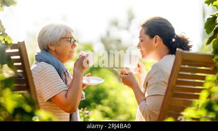 Happy young woman and her mother drinking tea in summer morning. Family sitting in the garden with cups and enjoying the conversation. Stock Photo