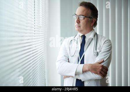 Pensive physician standing at medical office window and looking away Stock Photo