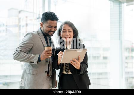 Two professional Asian male and female executive managers are looking at a tablet screen and discussing work while standing in the office building tog Stock Photo