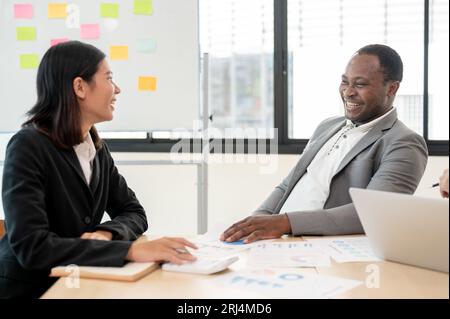 A satisfied and happy African American businessman or male CEO is laughing and enjoying discussing work with an Asian female assistant in the meeting. Stock Photo
