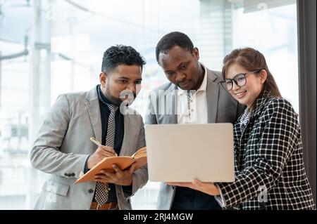 A multiracial group of business people is looking at a laptop screen and discussing work while standing in a company corridor together. diverse collea Stock Photo