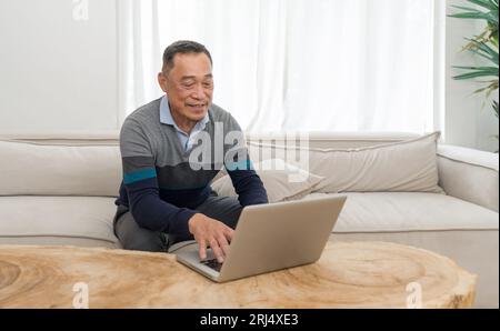 Home office scenario capturing senior man comfortably sitting on a couch, busy working or surfing the internet on his laptop. Stock Photo