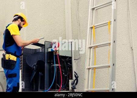 Precise worker commissioned for external condenser annual check up, writing report on clipboard. Licensed technician doing hvac unit safety check, looking for system electrical issues Stock Photo