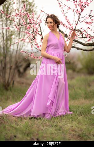 Woman peach blossom. Happy curly woman in pink dress walking in the garden of blossoming peach trees in spring. Stock Photo