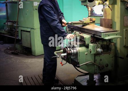 A male worker works on a larger metal iron locksmith lathe, equipment for repairs, metal work in a workshop at a metallurgical plant in a repair produ Stock Photo