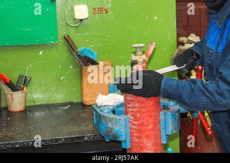 A male worker on a large metal industrial vice is repairing a red fire extinguisher cylinder in a workshop at the plant. Stock Photo