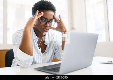 Puzzled by difficult task female programmer thinking about resolving. Concentrated african-american female office employee staring at laptop screen, deep in thoughts Stock Photo