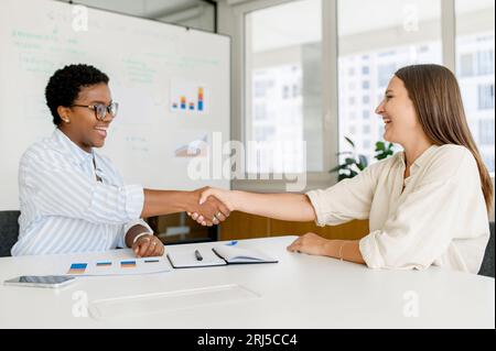 Two businesswomen sitting at the desk and shaking hands each other in sign of cooperation. Diverse female office employees greeting each other. Business partners made a deal Stock Photo