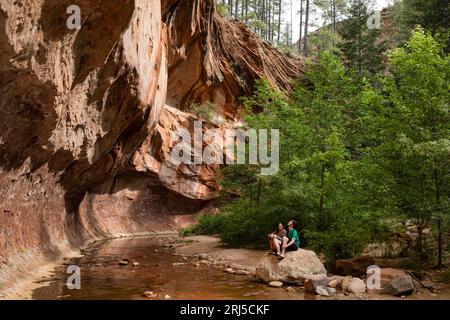 Young couple sit on rock by stream under rock ledge Stock Photo