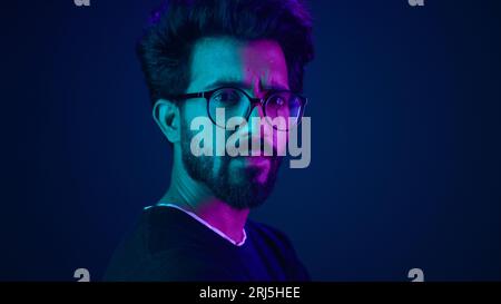Indian man guy profile turning head looking camera serious developer coding internet technology hacker programmer Arabian male in glasses neon blue Stock Photo