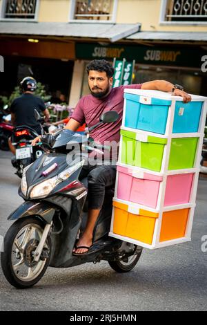 A guy takes a huge risk travelling down Soi Buakhao, Pattaya on his motorbike whilst carrying a large plastic cabinet. Stock Photo