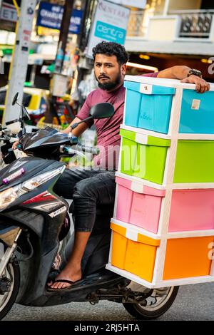 A guy takes a huge risk travelling down Soi Buakhao, Pattaya on his motorbike whilst carrying a large plastic cabinet. Stock Photo