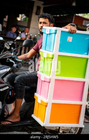 A guy takes a huge risk travelling down Soi Buakhao, Pattaya on his motorbike whilst carrying a large plastic cabinet. Stock Photo