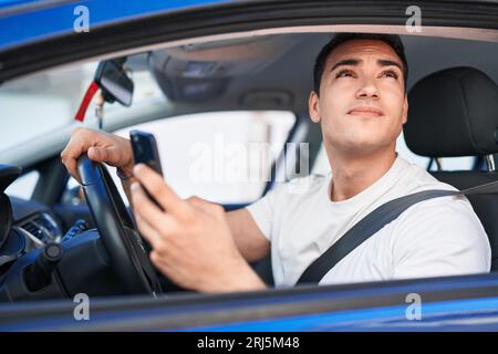 Young hispanic man using smartphone sitting on car at street Stock Photo