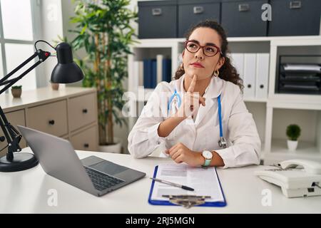 Young hispanic woman wearing doctor uniform and stethoscope thinking concentrated about doubt with finger on chin and looking up wondering Stock Photo