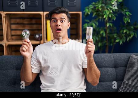 Hispanic man holding led lightbulb and incandescent bulb in shock face, looking skeptical and sarcastic, surprised with open mouth Stock Photo