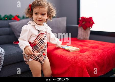 Adorable hispanic girl playing xylophone standing by christmas decoration at home Stock Photo