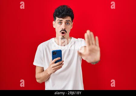 Hispanic man using smartphone over red background doing stop gesture with hands palms, angry and frustration expression Stock Photo