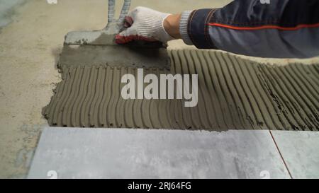 A man puts tiles on the wall. Builder, tiler puts gray ceramic tiles on the wall in the bathroom. Applying adhesive solution on the wall, for gluing Stock Photo