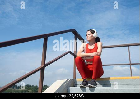 female athlete in sportswear wearing wireless headphones and listening to music, sitting on steps resting. Outdoor fitness concept. Stock Photo