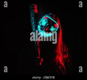 A crazy girl in an ominous neon mask holding a wired baseball bat on her shoulder, posing in a dark room Stock Photo