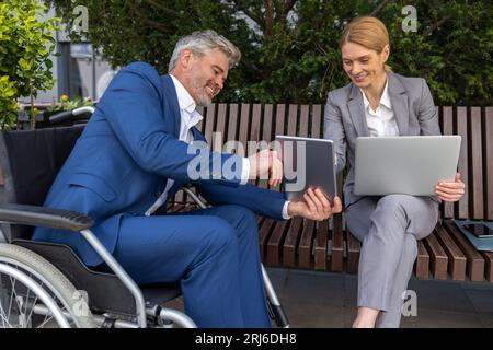 Businessman in wheelchair with female partner using laptop and tablet for working on joint project Stock Photo