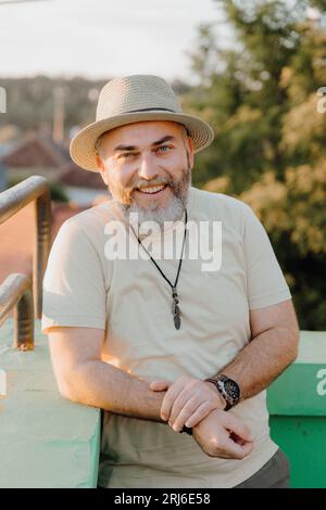 A mature man wearing a hat is sitting in a relaxed pose, facing the camera and smiling Stock Photo