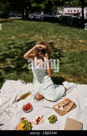 african american woman taking photo on vintage camera near wine and strawberries, summer picnic Stock Photo