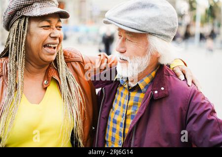 Multiracial senior couple having fun outdoor during fall time - Asian mature man and african woman laughing together while walking on city streets - J Stock Photo