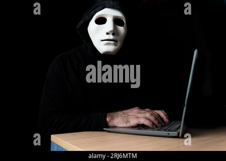 hacker dressed in black with mask and hood typing on a computer while looking at the camera cybercrime concept Stock Photo