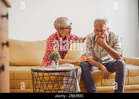 Senior couple sitting in living room. Man is sad and worried and woman is consoling him. Stock Photo