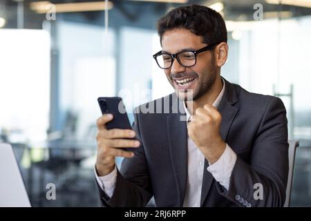 Close-up portrait of successful businessman, winner holding phone, man celebrating good results achievement received good news online, using app on smartphone inside office. Stock Photo