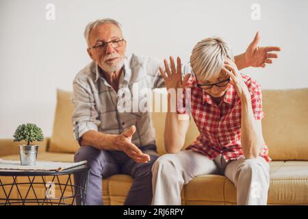 Senior couple sitting in living room and arguing. Woman is angry while man is apologizing. Stock Photo
