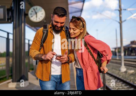 Happy couple is standing at railway station and waiting for arrival of their train. They are looking at map on smartphone. Stock Photo