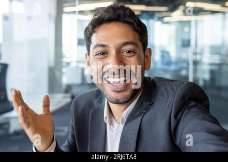 Video call friendly conversation, arab businessman inside office talking with friends and colleagues at workplace, man using app on phone, smiling and friendly waving at camera. Stock Photo