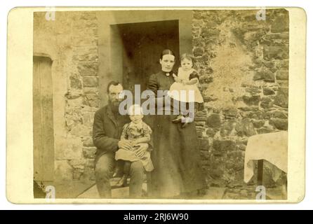 Original 1890's era charming Victorian cabinet card of a farming Victorian family outside their rustic stone farmhouse or cottage. The mother wearing her Sunday best, neat hairstyle with centre parting, holds her baby / toddler girl, the father, bearded, wearing a sack coat,  sits a child on his knee, who looks like a boy although wearing a girl's dress as was normal at the time. U.K. circa 1890,1891 Stock Photo