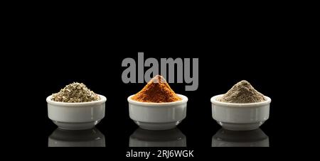 Three white porcelain cups with different spices - oregano, black and red pepper stand on a black mirror background and are beautifully reflected in i Stock Photo