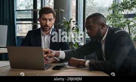 Two diverse multiracial businessmen discuss online data project with laptop in office writing notes documents. Caucasian businessman and African Stock Photo