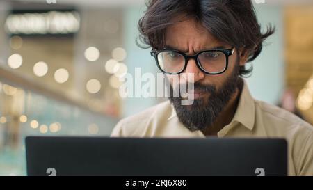 Serious Indian man in glasses looking at laptop screen working online indoors. Close up Arabian businessman in eyeglasses chatting with computer Stock Photo
