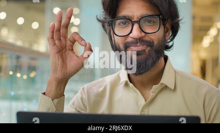 Indian Arabian bearded business man entrepreneur in glasses typing on laptop male professional businessman using computer sitting at office showing ok Stock Photo