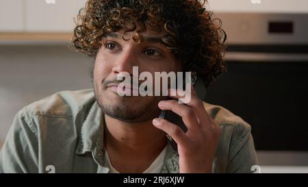 Cheerful Indian young man with curly hair talking phone in home kitchen. Happy friendly Hispanic guy talk with friends by smartphone Arabian male Stock Photo