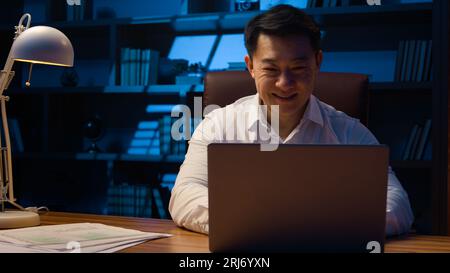 Middle-aged happy smiling businessman working with laptop and documents at dark night office Asian Korean man financial analyst accounting business Stock Photo