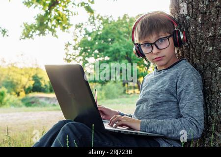 Boy with inquisitive blue eyes in glasses studying while sitting by tree with laptop on lap, listening to task through headphones. Student, surprised Stock Photo